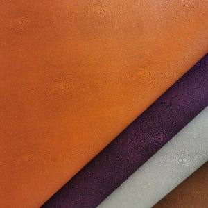 May include: Close-up of several rolls of textured leather in various colors. The colors include orange, purple, gray, and brown. The leather has a bumpy texture with small, evenly spaced dots.