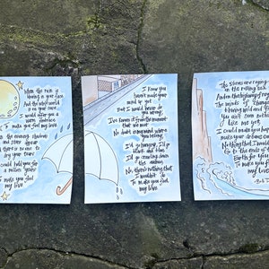 May include: Three watercolor art prints with song lyrics. The first features a moon and stars, the second an umbrella and street scene, and the third waves. Each print has handwritten text from Bob Dylan's song, "To Make You Feel My Love."