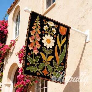 May include: A black garden flag with a floral design featuring orange, white, and green flowers. The flag has a geometric border and is mounted on a white pole. It hangs outside a building with pink flowers, adding a touch of colour.