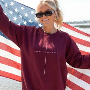 May include: A maroon long-sleeve crewneck sweatshirt featuring the text "ONE NATION UNDER GOD" printed in white above a thin vertical white line. The sweatshirt is worn in front of a large American flag with red, white, and blue stripes and stars.