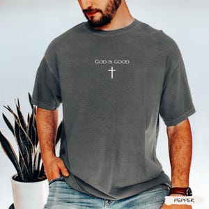 Peut inclure: Un t-shirt gris avec le texte "GOD IS GOOD" et une croix blanche. Le t-shirt a des manches courtes et un col rond. Le t-shirt est fait d'une mati&egrave;re douce.