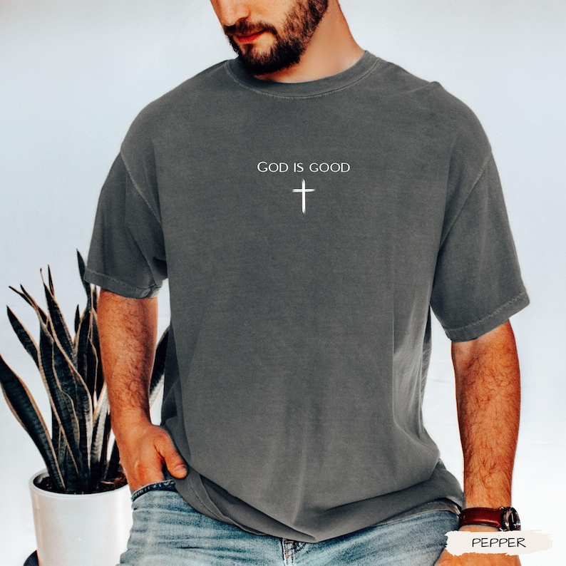 Peut inclure: Un t-shirt gris avec le texte "GOD IS GOOD" et une croix blanche. Le t-shirt a des manches courtes et un col rond. Le t-shirt est fait d'une mati&egrave;re douce.