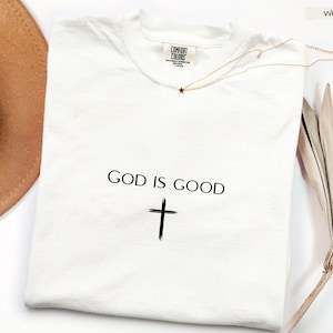 Peut inclure: T-shirt blanc avec l'inscription "GOD IS GOOD" et une croix noire. Un collier dor&eacute; avec un pendentif en forme d'&eacute;toile est pos&eacute; sur le t-shirt. Un chapeau marron et une plante s&eacute;ch&eacute;e sont en arri&egrave;re-plan.