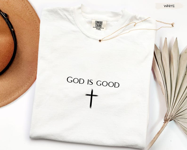 Peut inclure: T-shirt blanc avec l'inscription "GOD IS GOOD" et une croix noire. Un collier dor&eacute; avec un pendentif en forme d'&eacute;toile est pos&eacute; sur le t-shirt. Un chapeau marron et une plante s&eacute;ch&eacute;e sont en arri&egrave;re-plan.