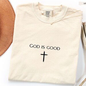 Peut inclure: T-shirt Comfort Colors de couleur ivoire avec l'inscription "GOD IS GOOD" et une croix noire. Le t-shirt est pr&eacute;sent&eacute; avec un chapeau marron et une branche de feuilles s&eacute;ch&eacute;es.