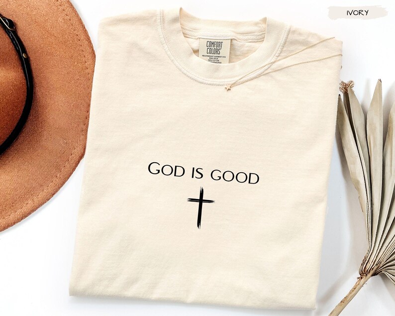 Peut inclure: T-shirt Comfort Colors de couleur ivoire avec l'inscription "GOD IS GOOD" et une croix noire. Le t-shirt est pr&eacute;sent&eacute; avec un chapeau marron et une branche de feuilles s&eacute;ch&eacute;es.