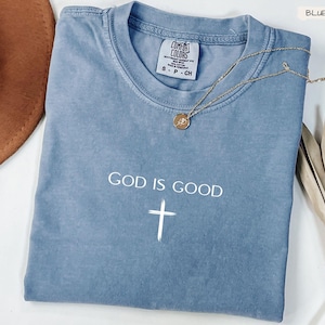Peut inclure: Un t-shirt bleu jean Comfort Colors avec les mots "GOD IS GOOD" et une croix blanche. Un collier en or avec un pendentif est drap&eacute; sur le t-shirt. Un chapeau marron et une plante s&eacute;ch&eacute;e sont en arri&egrave;re-plan.