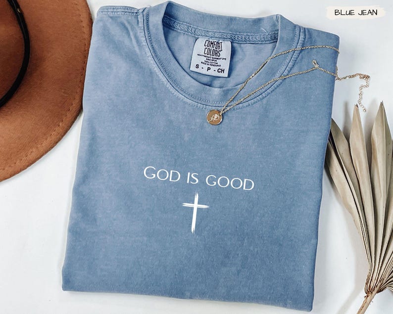 Peut inclure: Un t-shirt bleu jean Comfort Colors avec les mots "GOD IS GOOD" et une croix blanche. Un collier en or avec un pendentif est drap&eacute; sur le t-shirt. Un chapeau marron et une plante s&eacute;ch&eacute;e sont en arri&egrave;re-plan.