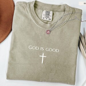 Peut inclure: T-shirt kaki avec l'inscription "GOD IS GOOD" et une croix blanche. Un collier argent&eacute; avec un pendentif est drap&eacute; sur le t-shirt. Le t-shirt porte une &eacute;tiquette Comfort Colors.