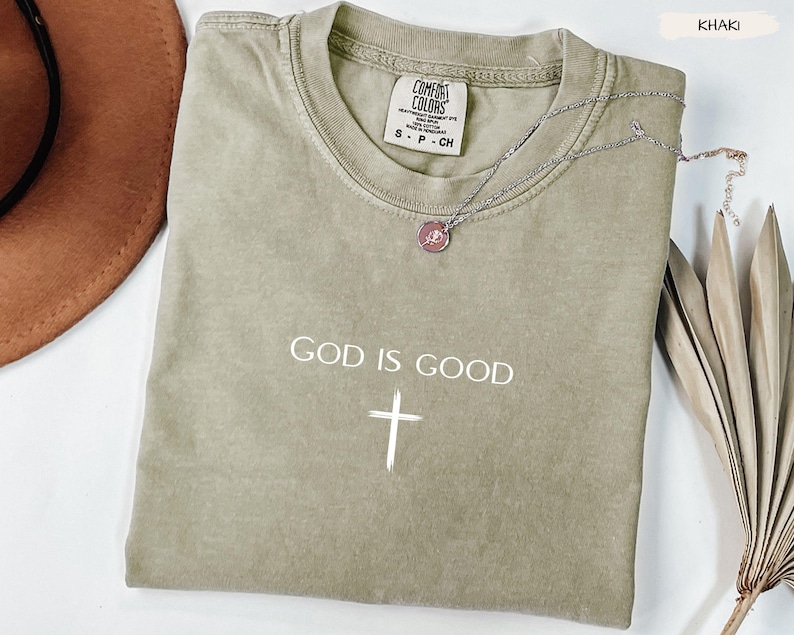 Peut inclure: T-shirt kaki avec l'inscription "GOD IS GOOD" et une croix blanche. Un collier argent&eacute; avec un pendentif est drap&eacute; sur le t-shirt. Le t-shirt porte une &eacute;tiquette Comfort Colors.
