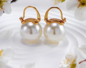 Puede incluir: Un par de pendientes de perlas con aros dorados. Los pendientes son redondos y tienen una superficie blanca y brillante. El fondo presenta flores y pétalos blancos, creando una estética delicada y elegante.