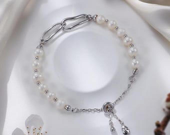 Puede incluir: Una delicada pulsera con perlas blancas y una cadena de plata. La pulsera tiene un diseño único con un eslabón alargado y un cierre deslizante. El fondo incluye flores blancas y una tela blanca sedosa.