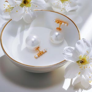 May include: Elegant pearl earrings with gold accents rest in a white bowl with a gold rim, alongside delicate white flowers. The earrings feature large, lustrous pearls and small gold bars with pearls. A sophisticated jewelry piece.