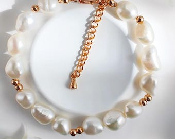 Puede incluir: Un brazalete de perlas blancas de forma irregular, con detalles dorados. El cierre y la cadena también son dorados. El brazalete se exhibe sobre una superficie blanca, con flores blancas en el fondo. Longitud ajustable.