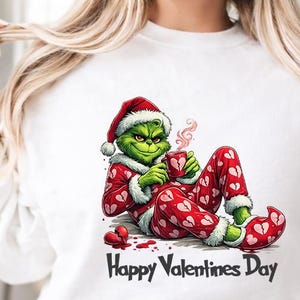 May include: White sweatshirt featuring a cartoon Grinch character in a Santa hat and pajamas with heart patterns, holding a mug. The text "Happy Valentines Day" is printed below the character.