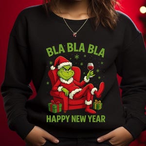May include: Black sweatshirt featuring a Grinch character in a Santa suit, holding a wine glass, and seated in a red chair. The text "BLA BLA BLA" and "HAPPY NEW YEAR" are in green. Two wrapped gifts are at the bottom.