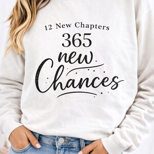 Pode incluir: Camisola creme com o texto preto "12 New Chapters 365 new Chances". A camisola é combinada com jeans azuis. A imagem foca-se na mensagem da camisola, sugerindo um tema de novos começos e oportunidades.