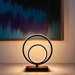 Pode incluir: Candeeiro de mesa LED moderno a preto e branco com design circular. O candeeiro apresenta dois anéis de luz concêntricos, com um círculo interior menor. O candeeiro assenta numa base retangular, em frente a uma pintura abstrata.