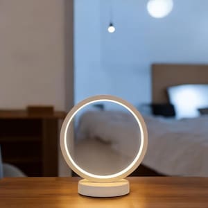 May include: A white circular table lamp with a ring of light around the edge. The lamp is on a wooden table and is illuminated.