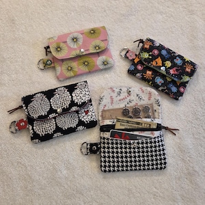 May include: Four small fabric wallets in different patterns. One pink wallet with yellow and white floral design, one black wallet with white sheep, one black wallet with colourful sea creatures, and one black and white houndstooth wallet.