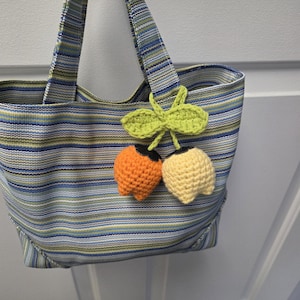 May include: A striped tote bag in blue, green, and yellow with a crochet cherry embellishment. The bag has a rectangular shape with handles. The crochet cherries are orange and yellow with green leaves.