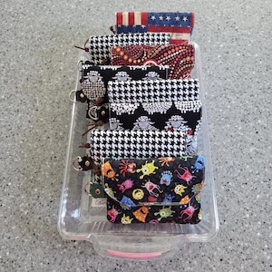 May include: A collection of small fabric pouches with snap closures, featuring various patterns including American flag, houndstooth, sheep, colourful monsters, and abstract dots, arranged in a clear rectangular plastic tray.
