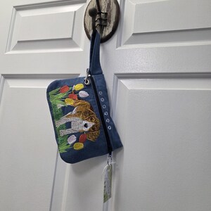 May include: A denim clutch bag with a zip and strap, suspended from a hook. It has an embroidered design of a dog in a field of red, yellow, and pink tulips. The bag is a practical accessory.