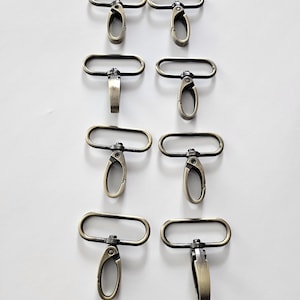 May include: Eight antique brass-colored swivel clasps with oval rings and lobster claw closures are arranged on a white background. The clasps are designed for attaching to bags, straps, or other accessories.