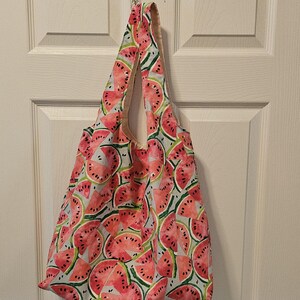 May include: A reusable tote bag featuring a watermelon print. The bag is made from lightweight fabric and has a single handle.