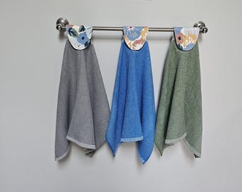 May include: Three decorative hand towels hanging on a metal bar. Each towel has a floral patterned top and a solid-colored bottom in gray, blue, and green. The towels are made of a textured fabric.