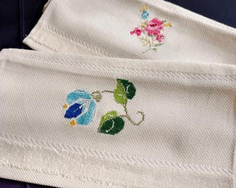Puede incluir: Dos toallas de mano de lino color crema con diseños florales bordados. Una toalla presenta una flor azul con hojas verdes, mientras que la otra tiene un ramo de flores rosas y azules. Las toallas tienen un tejido texturizado y un borde cosido decorativo.