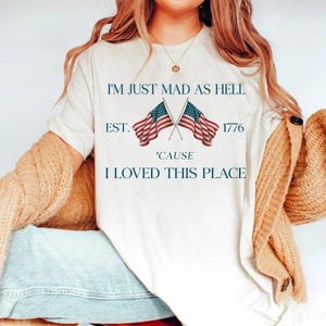 May include: Cream-colored t-shirt with the text "I'M JUST MAD AS HELL EST. 1776 'CAUSE I LOVED THIS PLACE" and two crossed American flags. The shirt is a casual style.