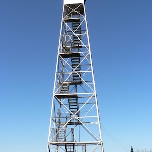 May include: A tall, white metal fire lookout tower with a square observation cabin at the top. The structure features a series of open-air staircases and landings arranged in a zig-zag pattern within the framework, set against a clear blue sky.
