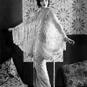 May include: A woman in a beaded, sequined gown with a flowing cape, standing in front of a patterned wall. She is wearing a simple hairstyle and has a neutral expression.