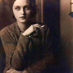 May include: A woman wearing a dark beret and a brown patterned jacket, with a cigarette in her hand, poses for a portrait.