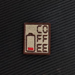 Puede incluir: Un parche cuadrado con la palabra "COFFEE" dispuesta verticalmente. El parche presenta un icono de batería con un indicador de carga baja. El parche es beige con costuras marrones y un borde marrón, sobre un fondo negro texturizado.