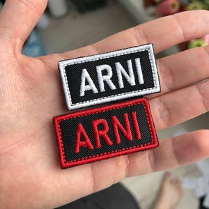 Puede incluir: Dos parches rectangulares bordados con el nombre "ARNI". Un parche tiene texto blanco sobre fondo negro con borde blanco. El otro tiene texto rojo sobre fondo negro con borde rojo. Los parches se sostienen en una mano.