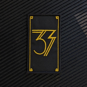 Puede incluir: Un parche rectangular negro bordado con un borde amarillo y detalles decorativos en las esquinas. En el centro aparece el número 37 bordado en amarillo, donde el siete tiene un diseño angular de rayo.
