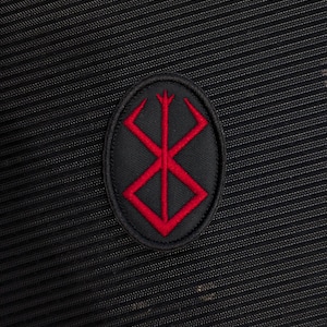 May include: An oval-shaped embroidered patch featuring a red symbol of a stylized brand on a black background. The patch is stitched onto a black fabric surface with a fine, ribbed texture that runs diagonally across the entire frame.