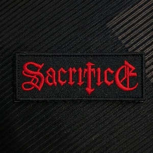 Puede incluir: Un parche rectangular bordado en negro con la palabra Sacrifice cosida en una fuente gótica de color rojo intenso. El parche tiene un borde negro definido y una base de tela texturizada, diseñado para su aplicación en ropa o textiles.