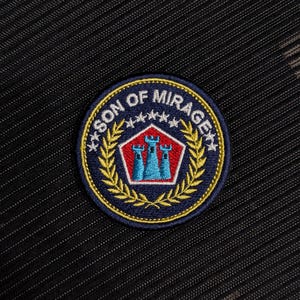 May include: Circular embroidered patch with the text "SON OF MIRAGE" in white and gold. The patch features a red and blue castle design, surrounded by a gold laurel wreath, on a navy blue background. The patch is on a black textured fabric.