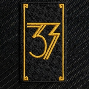 Könnte beinhalten: Ein rechteckiger bestickter Aufnäher mit schwarzem Hintergrund, goldenem Rand und der Zahl 37 in einer stilisierten goldenen Schrift. Das Design umfasst dekorative Eckakzente und eine saubere, strukturierte Verarbeitung auf schwarzem Stoff.