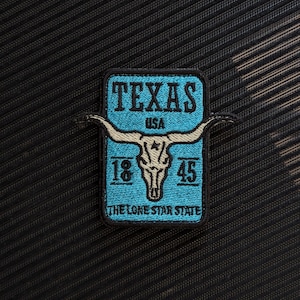 May include: Embroidered patch with a turquoise background, displaying "TEXAS" in bold letters. A steer skull graphic is centered, flanked by "18" and "45". The text "THE LONE STAR STATE" is at the bottom. The patch is on a textured black surface.