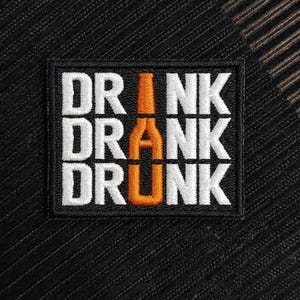 Pode incluir: Um emblema bordado retangular com borda e fundo pretos. O texto DRINK DRANK DRUNK é bordado em branco, com um ícone de garrafa laranja integrado no centro de cada palavra, substituindo as letras I, A e U, respectivamente.
