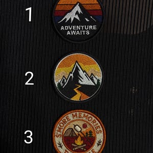 Op de afbeelding: Drie ronde geborduurde patches met outdoorthema's. De bovenste patch heeft een bergontwerp met de tekst "ADVENTURE AWAITS". De middelste patch heeft een berg en een pad. De onderste patch zegt "S'MORE MEMORIES TO MAKE" met een kampvuur.