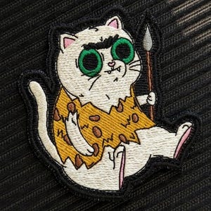 May include: An embroidered patch featuring a white cat with large green eyes and thick black eyebrows. The cat wears a yellow tunic with brown spots and holds a brown spear with a gray tip. The design is outlined in a thick black border.