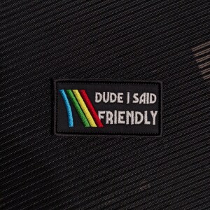 May include: Black fabric with a textured pattern. A rectangular patch is sewn onto the fabric, featuring the text "DUDE I SAID FRIENDLY" in white, with a colorful rainbow design.