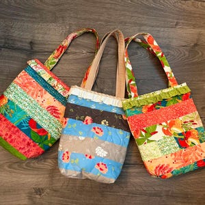 May include: Three quilted tote bags with fabric handles. The left bag features horizontal stripes of floral and green patterns. The center bag has blue, grey, and brown floral stripes with a white ruffle. The right bag has floral and green stripes.