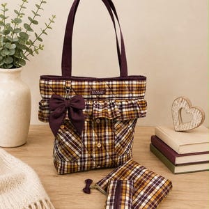 May include: A quilted tote bag featuring a brown, yellow, and white plaid pattern with a dark purple ruffled trim, a matching bow, and two shoulder straps. The bag displays a label reading "Austin Manor" and includes a smaller matching pouch.