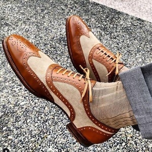 Può includere: Un paio di scarpe stringate brogue marroni e beige con intricate perforazioni e dettagli. Le scarpe presentano un design bicolore con pannelli in pelle e camoscio e sono allacciate con lacci color cuoio. Le scarpe sono indossate con calzini beige.